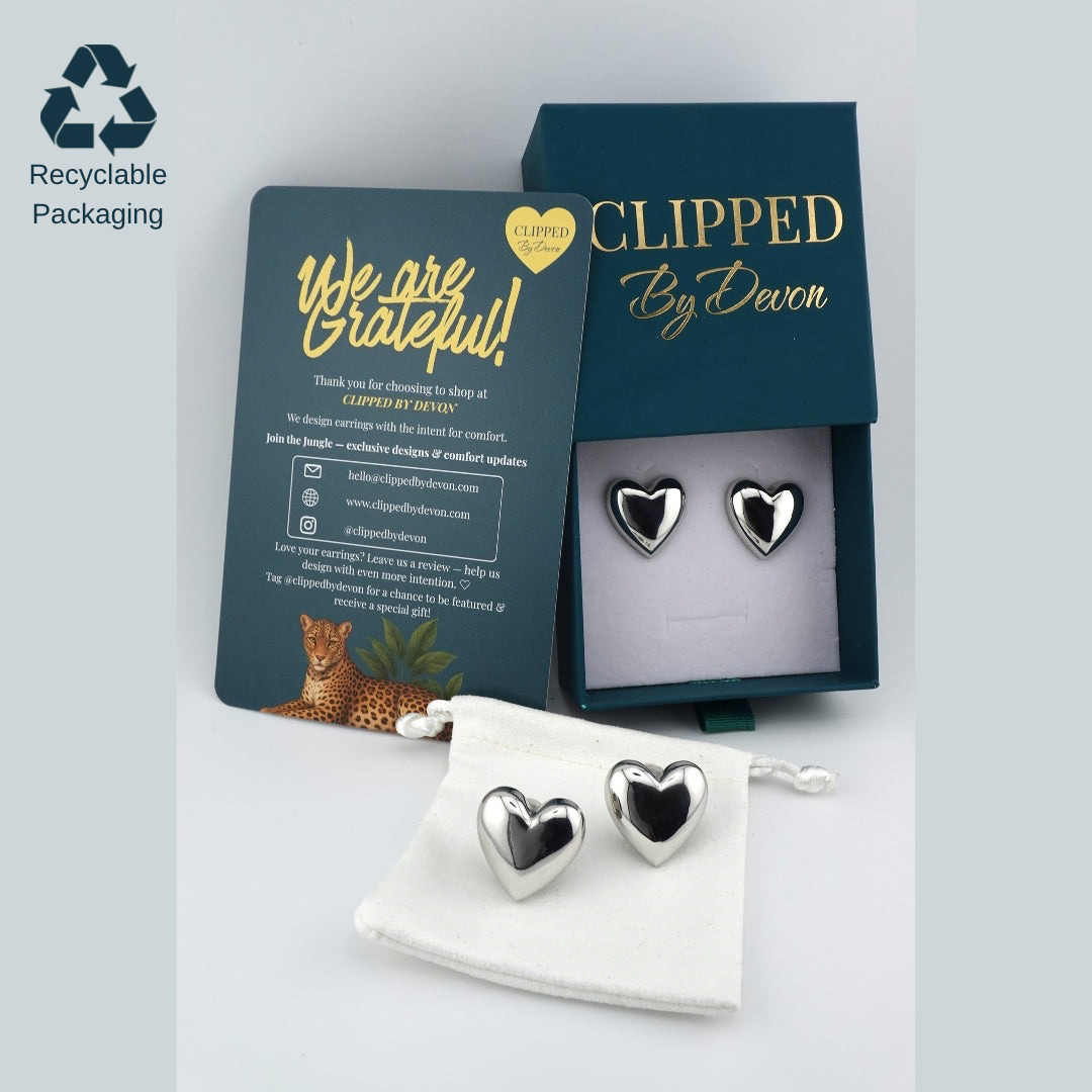 Silver heart clip-on earrings by Clipped by Devon displayed with recyclable jewelry box, thank-you card, and reusable pouch.